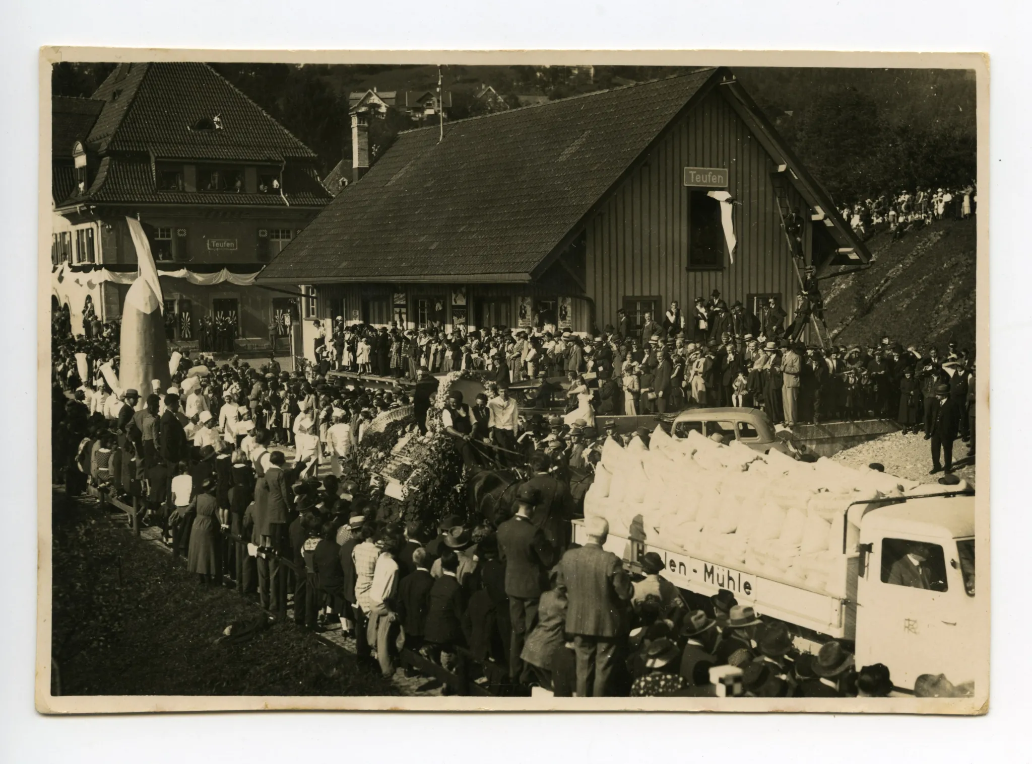 3 Wagen hintereinander: Lastwagen der Haslen-Mühle?, Fuhrwerk der Obst- und Gemüsebauern, Konditoren mit ihrer „Riesenpraline“, im Hintergrund Güterschuppen und Bahnhof