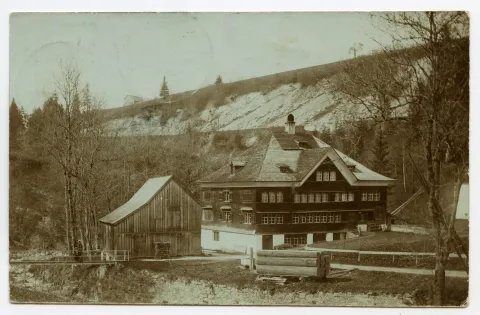 Ansicht mit einem Nebengebäude und aufgestapelten Holzplanken aus Süden, Personen auf dem Steg über den Rotbach