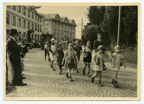 Gruppe von Kindern (Schüler)auf der Hauptstrasse gehend, Kinder mit Chränzen und Beerenkörgen, im Hintergrund Druckerei Kunz